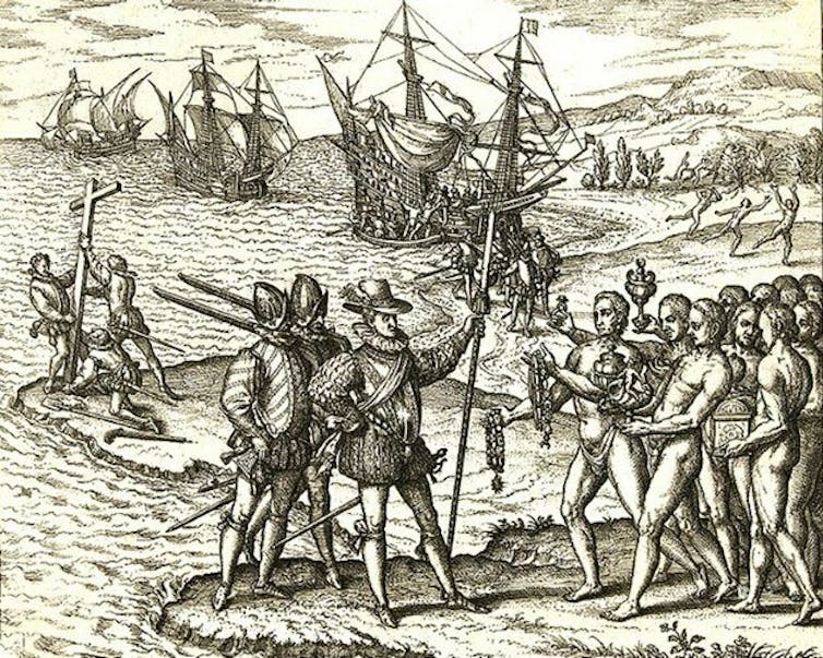 A black and white engraving of a group of armed and armored men standing on the shore speaking to many naked men. Large ships sail in the background.