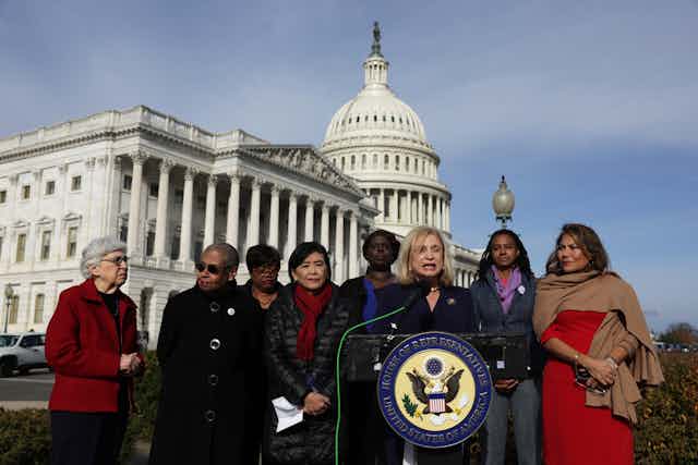 A group of women wear jackets and stand in front of a podium outside of the US Capitol. 