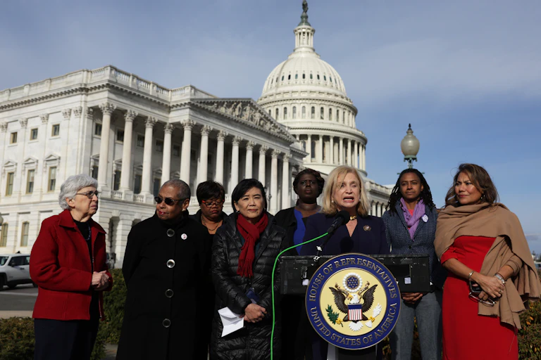 A group of women wear jackets and stand in front of a podium outside of the US Capitol.
