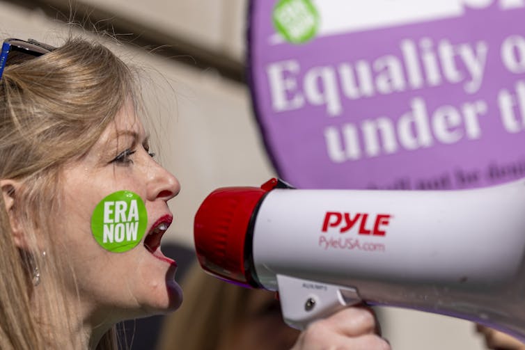 A blond woman yells into a megaphone and has a green sticker on her cheek that says 'ERA Now'