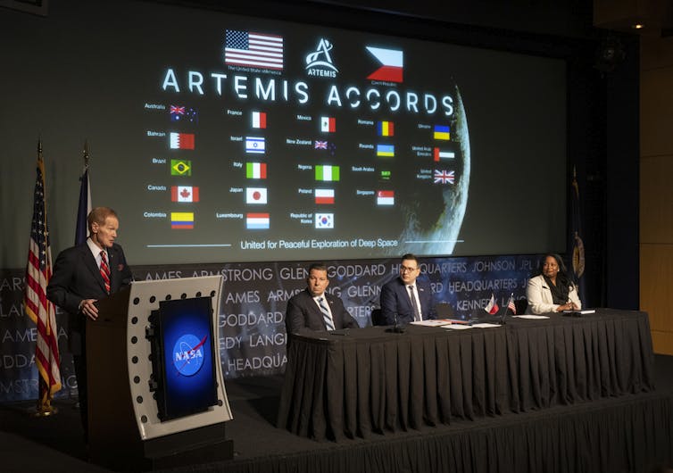 A suited man stands at a NASA podium, with three panelists seated at a table next to him. In the background a green reads 