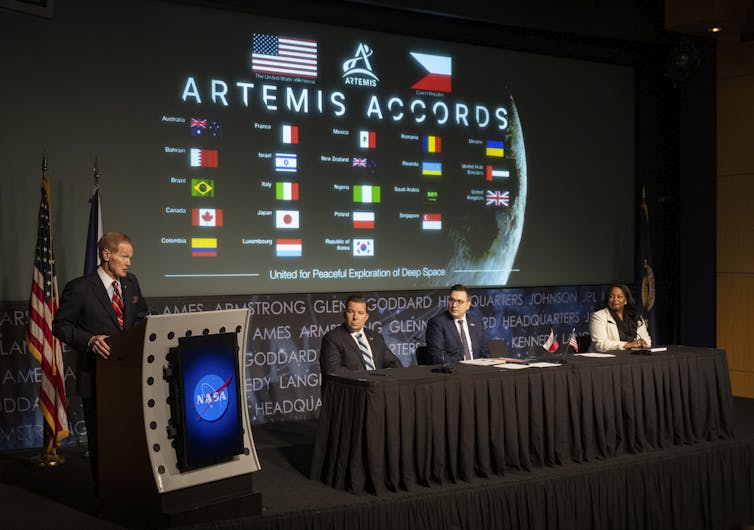A suited man stands at a NASA podium, with three panelists seated at a table next to him. In the background a green reads