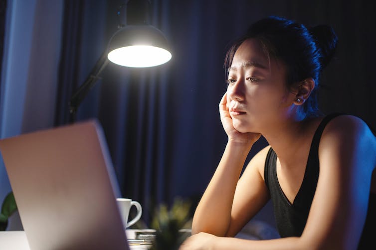 A tired woman is looking at a laptop with her chin in her hand.
