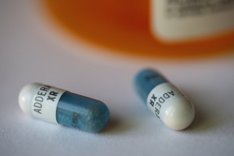 Two blue and white Adderall capsules lie the in the foreground with a medicine bottle sitting behind them.