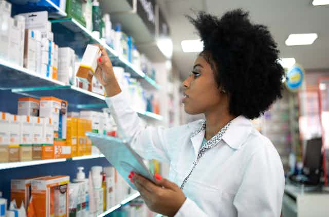 A female pharmacist holds up and inspects a box containing a drug in front of pharmacy shelves.