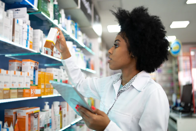 A female pharmacist holds up and inspects a box containing a drug in front of pharmacy shelves.