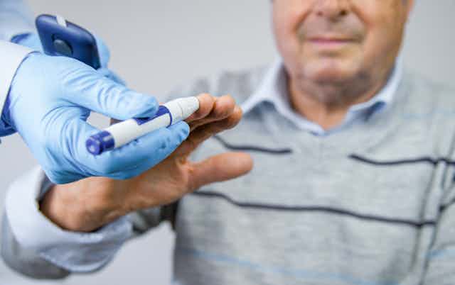 The gloved hand of a doctor or nurse holds a glucose meter up to the fingers of an elderly man to check his blood sugar levels. 