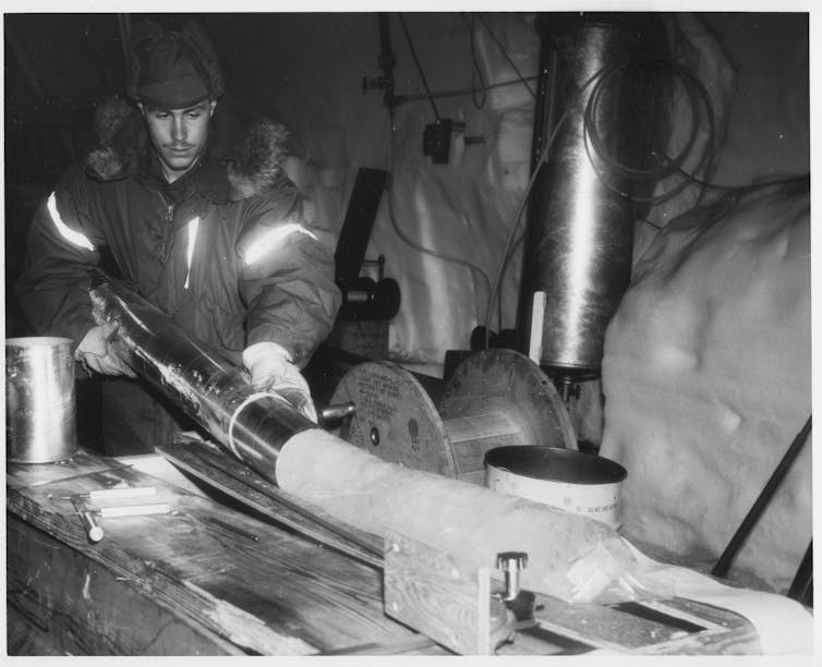 A man in a fur-lined coat removes a long ice core about as wide as his hand
