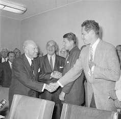 Men in suits, including Ronald Reagan, shake hands with one another in an old photo.