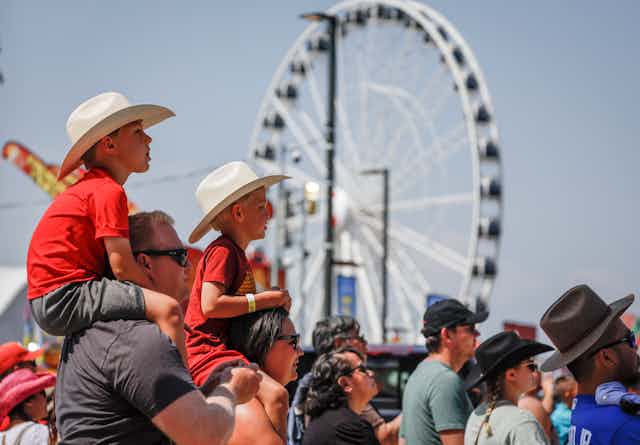 Two boys wearing cowboy hats while sitting on their parents' shoulders; a ferris wheel is in the background.