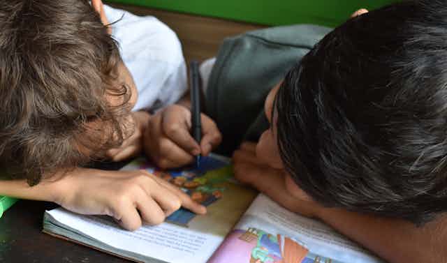 Two children seen huddled over a book, one holding a pen.