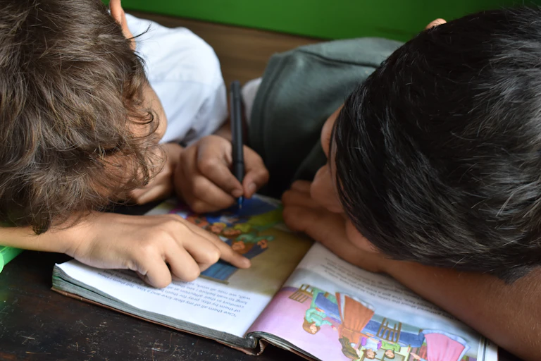Two children seen huddled over a book, one holding a pen.