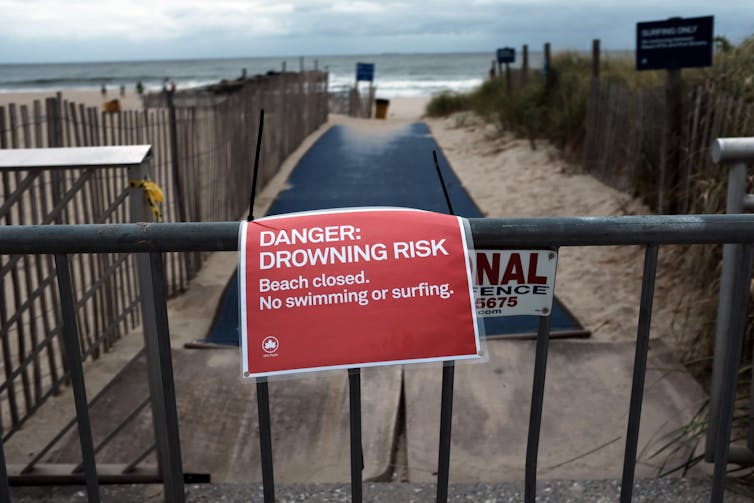 A barricaded pathway to a beach with a sign warning of drowning risk and barring swimming and surfing.