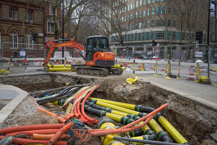 Roadworks with a digger at the side of a large hole with many water, gas and electricity pipes on display.