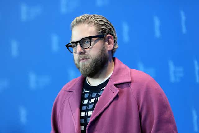 Jonah Hill posing on a red carpet in a pink coat. He wears black glasses and his hair is slicked back. 