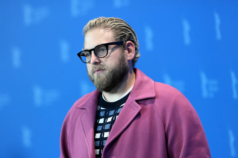 Jonah Hill posing on a red carpet in a pink coat. He wears black glasses and his hair is slicked back.