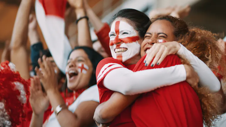 English female soccer fans with England flag painted on their faces hugging each other after their team's victory.