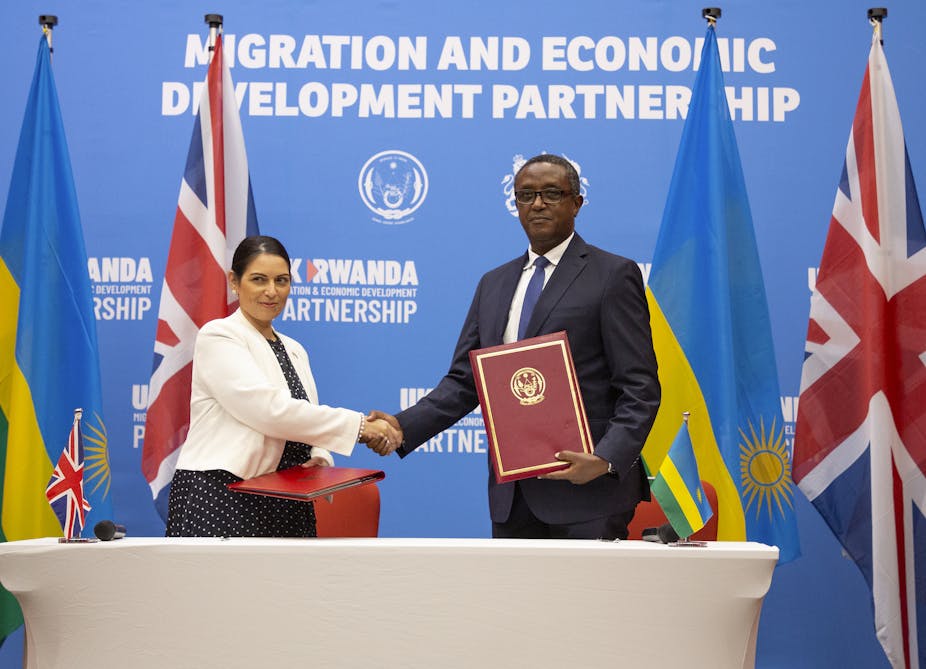 British Home Secretary Priti Patel (L) with Rwandan Foreign Minister Vincent Biruta shake hands in front of a blue wall reading Migration and Economic Development Partnership, and four Rwandan and UK flags.