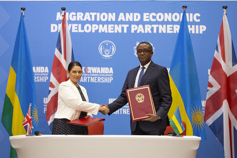 British Home Secretary Priti Patel (L) with Rwandan Foreign Minister Vincent Biruta shake hands in front of a blue wall reading Migration and Economic Development Partnership, and four Rwandan and UK flags.