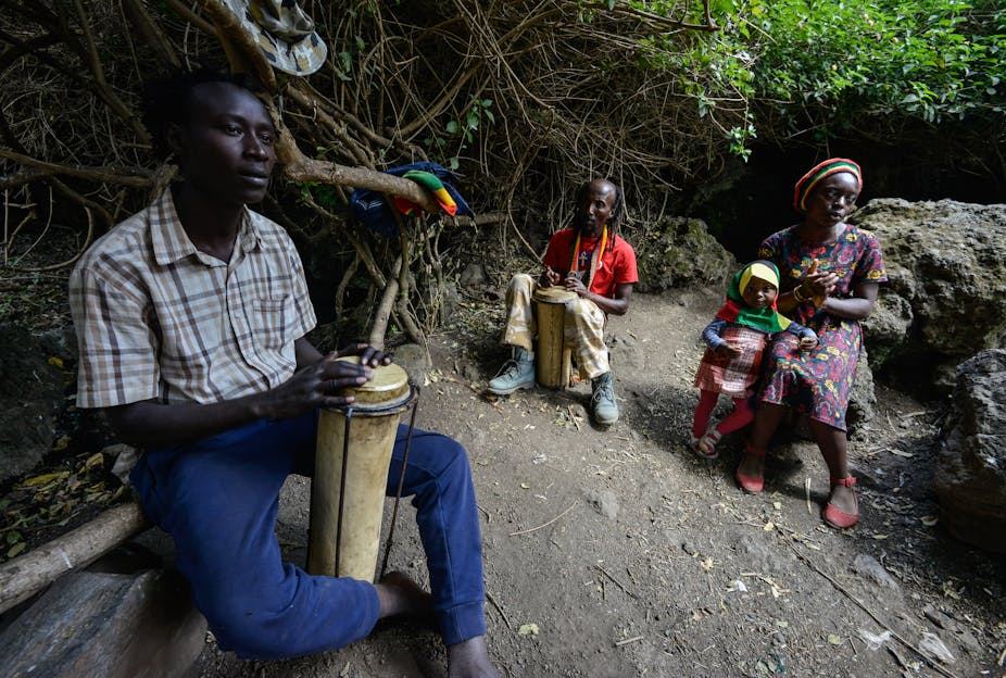 Two men with braided hair play a drum while a woman sits nearby on a rock, with a small child.