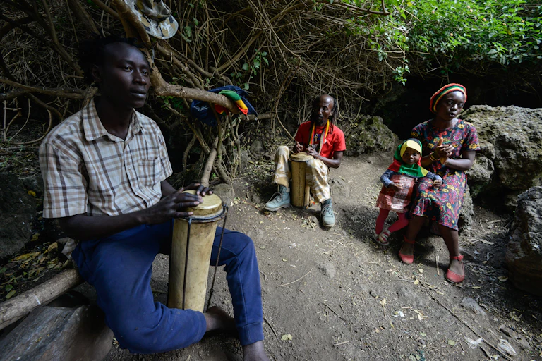 Two men with braided hair play a drum while a woman sits nearby on a rock, with a small child.