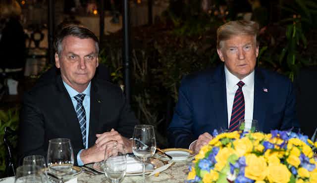 Two men in suits sit next to each other at a dinner table, place settings and a flower arrangement in front of them.