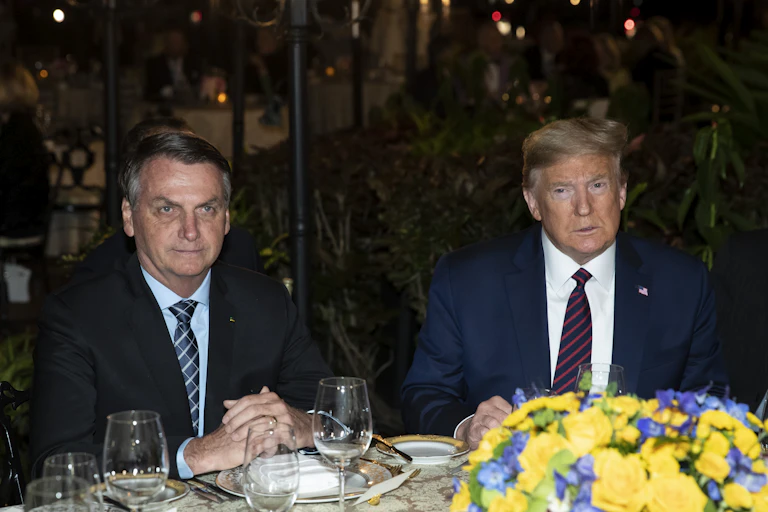 Two men in suits sit next to each other at a dinner table, place settings and a flower arrangement in front of them.