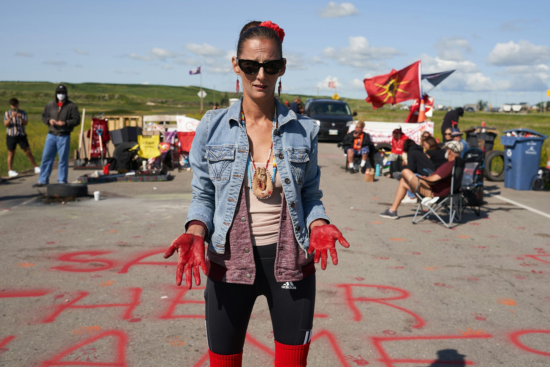 a woman standing on a road wearing a blue jacket and sunglasses poses for a photo with her palms covered in red paint.