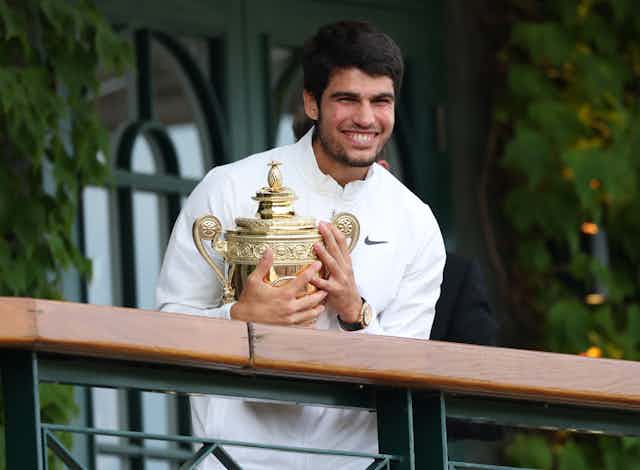 Carlos Alcaraz smiles in a white jumper, holding the gold Wimbledon trophy.