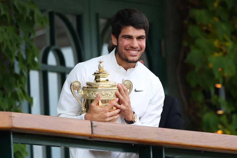 Carlos Alcaraz smiles in a white jumper, holding the gold Wimbledon trophy.