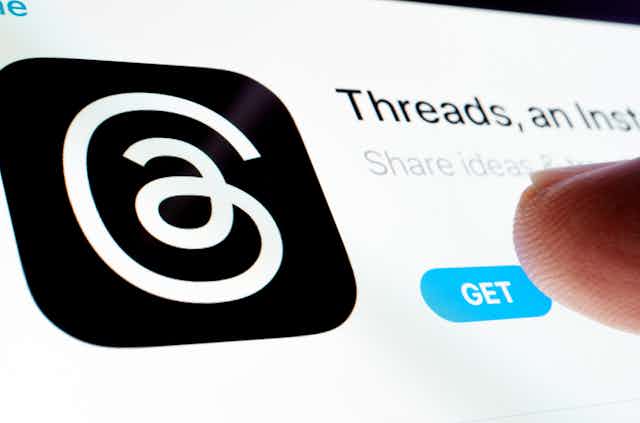 a close-up of a finger touching a screen showing the Threads logo, a curly white line on a black bacground