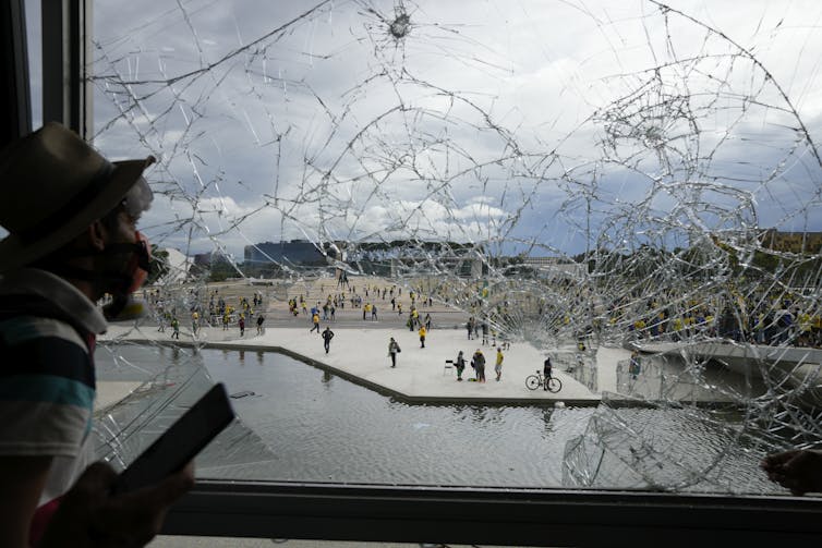 A man in a mask looks out a shattered window.