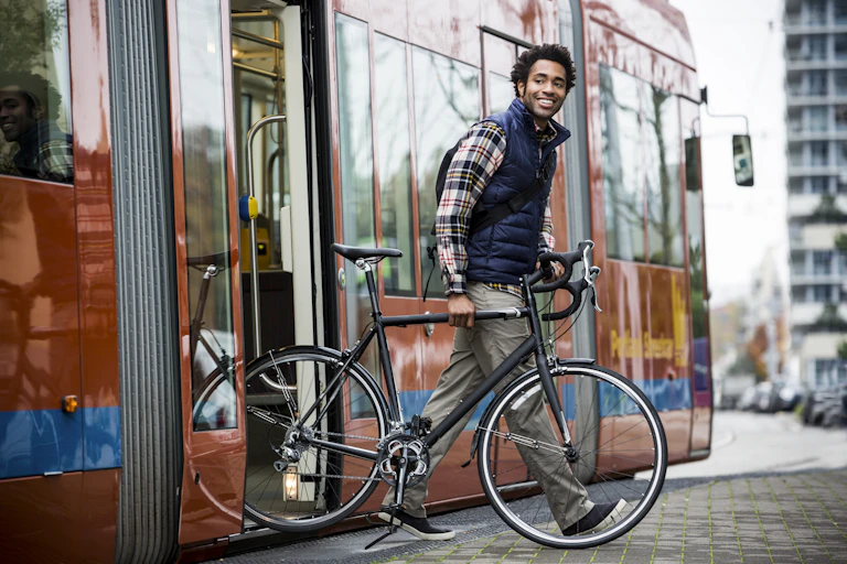 A many is walking off a train with a bike. He is smiling and wearing a longsleeve shirt and a vest.
