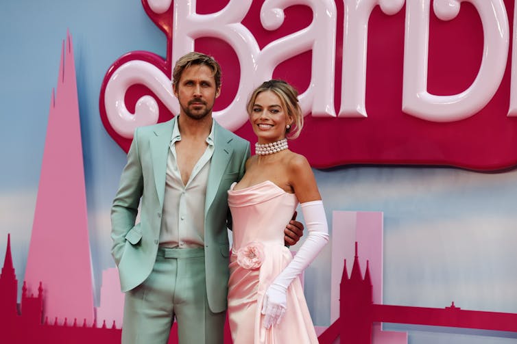 Ryan Gosling and Margot Robbie on the red carpet. Gosling wears a baby blue suit and Robbie a pink dress with white opera gloves.