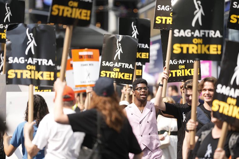 People holding protest signs that say 'SAG-AFTRA on Strike' walk down the sidewalk