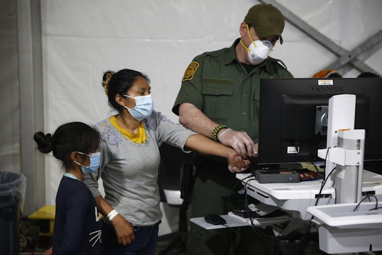 Una mujer usa una máscara facial y se para junto a una computadora junto a un niño pequeño.  Un hombre con uniforme verde y una máscara sostiene su dedo cerca de la computadora.