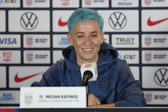 A woman with short, teal hair speaks into a microphone at a press conference