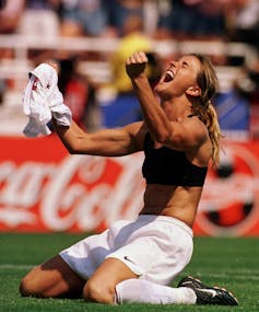 A woman in a sports bra and the lower half of a soccer uniform holds her shirt up with clenched fists as she yells in celebration