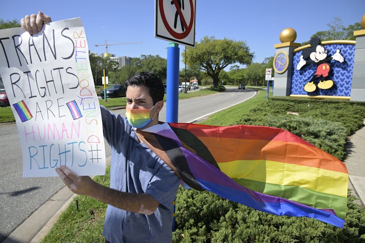 a man wearing a rainbow flag cape holds a pro-trans sign in front of a mickey logo