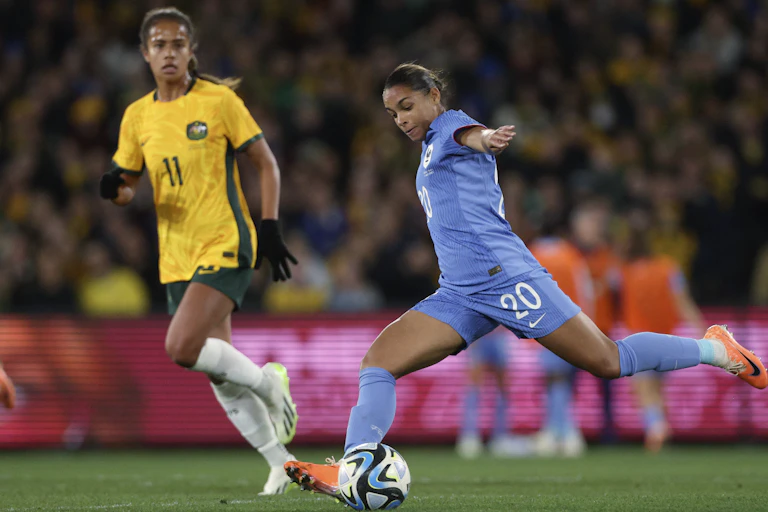 A woman in a blue soccer uniform kicks a ball while a woman in a yellow uniform looks on