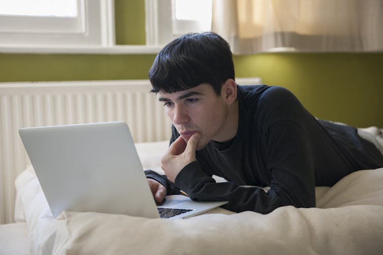 A teenager lies on his bed while looking at his laptop.