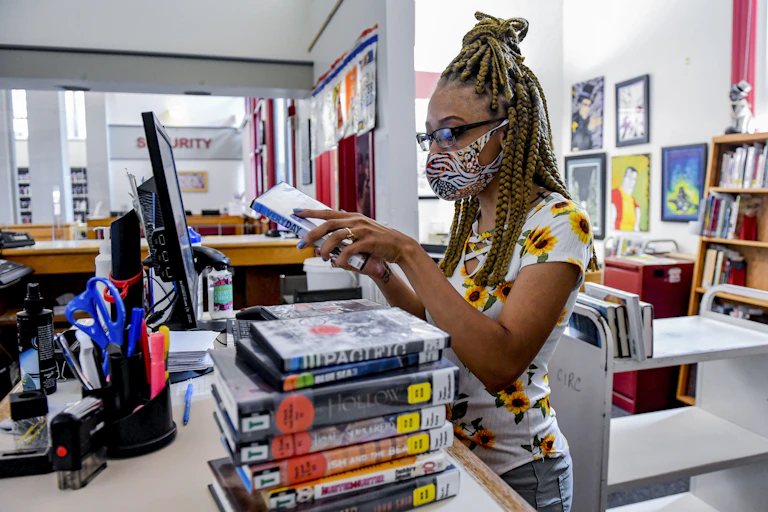 A woman in a white shirt with sunflowers on it stands at a table with a pile of books.