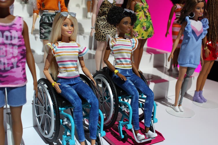 Fair skinned and dark skinned Barbies sitting in wheelchairs.