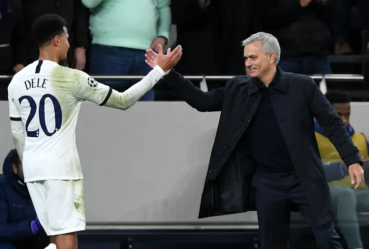 Dele and Jose Mourinho shaking hands, Dele wears a white football kit and Mourinho wears a black suit and coat.
