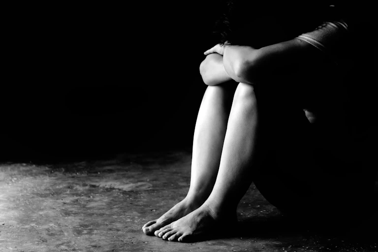 A black and white photo of a person sitting on the floor in the shadows grabbing their knees