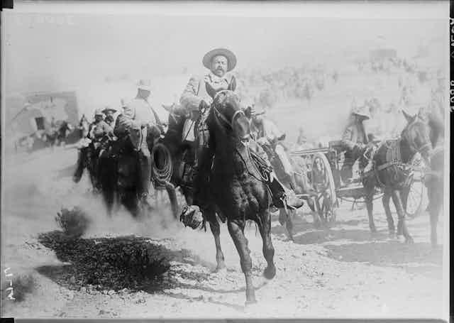 Un hombre con gorro mexicano monta a lomos de un caballo seguido por más hombres en caballo y carruajes.