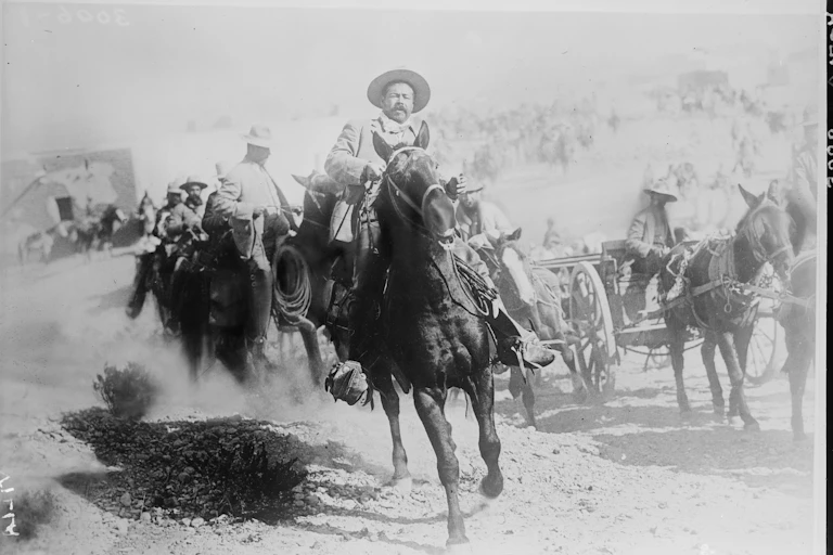 Un hombre con gorro mexicano monta a lomos de un caballo seguido por más hombres en caballo y carruajes.