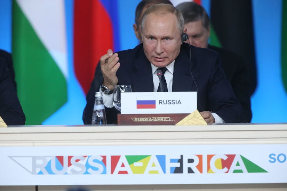 A man gesturing as he speaks into a microphone while seated. In front of him is a tag written 'Russia' and it sits on a table with the words 'Russia-Africa'