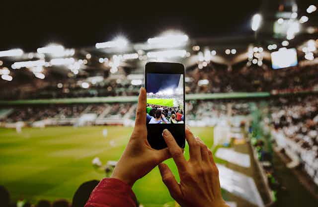 A woman's hands holding a phone to photograph a football pitch, from within the stadium seats.