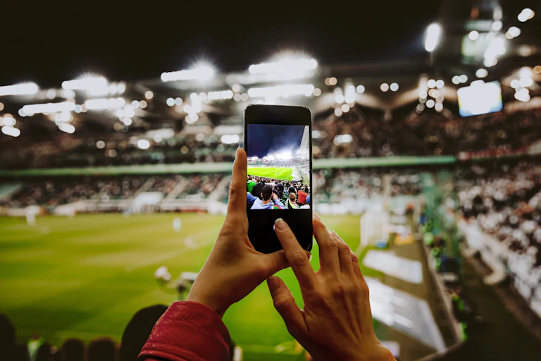 A woman's hands holding a phone to photograph a football pitch, from within the stadium seats.